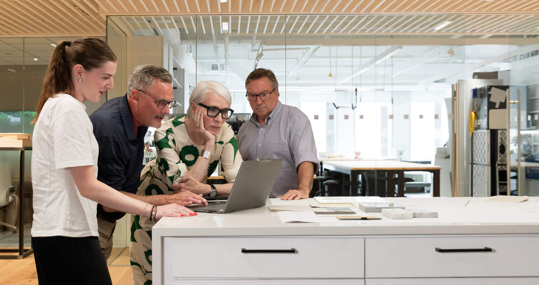 A group of four diverse professionals, focused on a laptop, collaborate in a bright workspace filled with design materials and models.