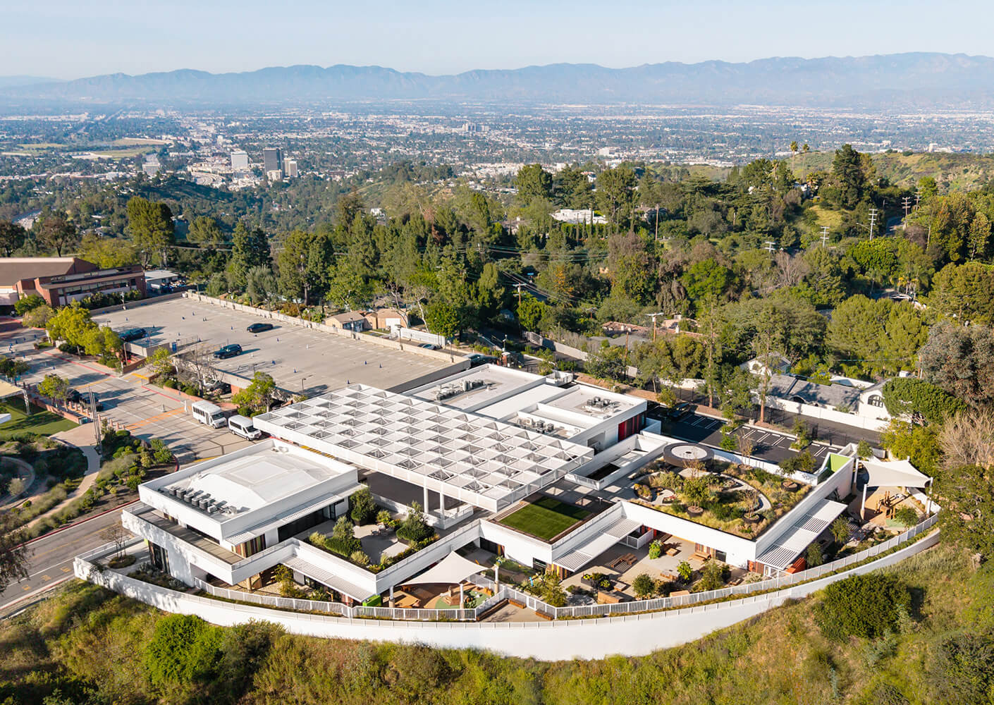 Aerial view of a multi-level building with landscaped outdoor spaces and a parking lot, surrounded by trees and hills.