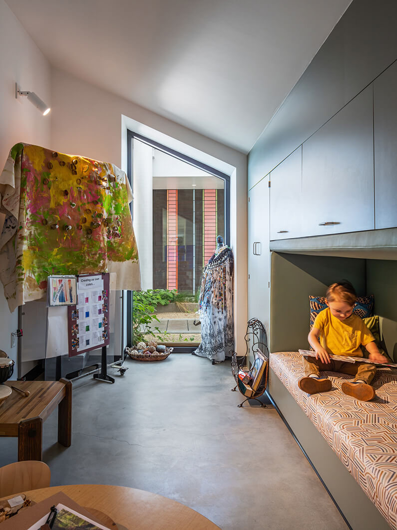 Child sitting on a patterned bench in a brightly lit room with artwork, plants, and a window showcasing an outdoor view.