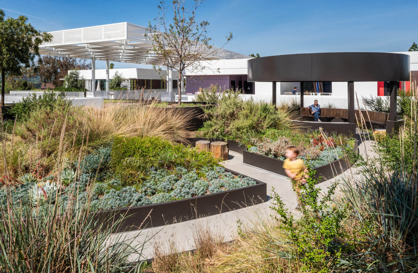 Landscape design featuring landscaped garden beds, seating areas, and children playing in an outdoor space with a clear blue sky.