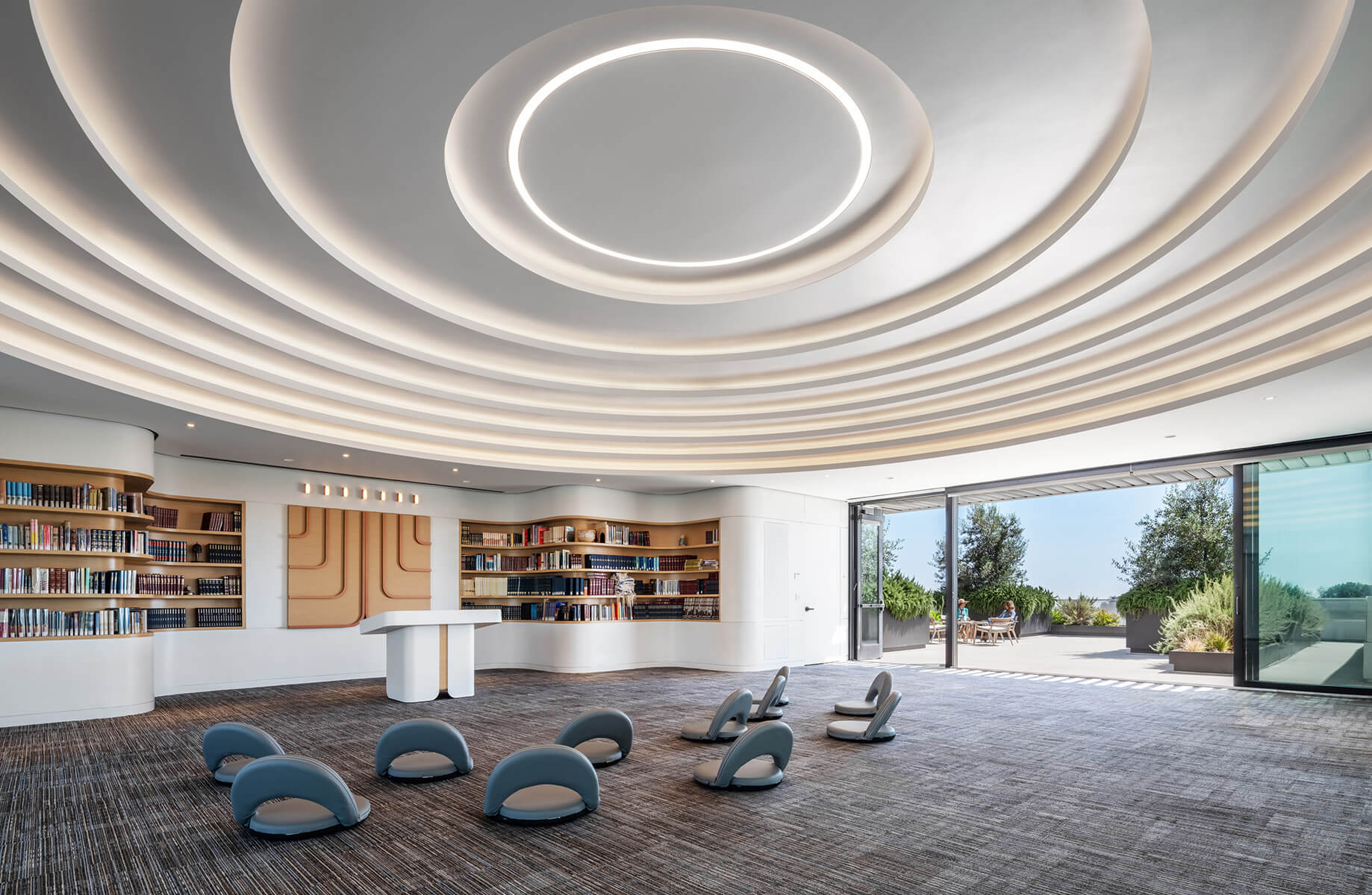 Interior view of a spacious library with curved ceiling, bookshelves, and seating. Large windows open to an outdoor area with greenery.