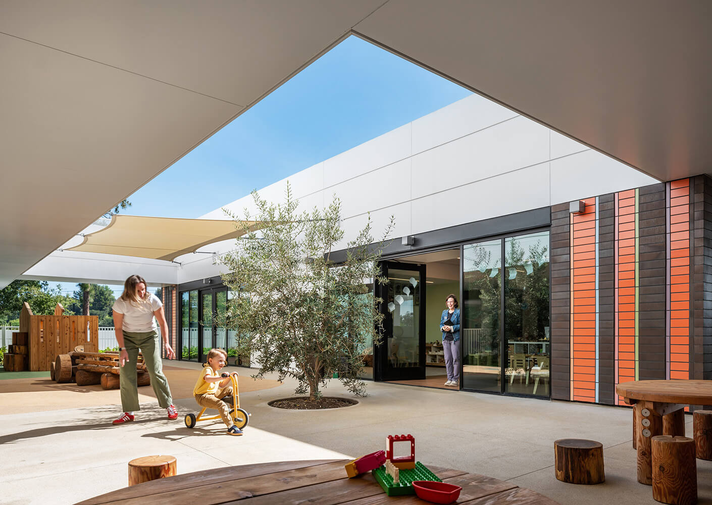 A caregiver and child interact outside a preschool, featuring a tree, shaded play area, and colorful building design. Toys are visible on the ground.