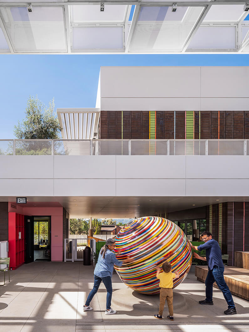 Children and adults interact with a large, colorful spherical sculpture in an outdoor architectural space, surrounded by modern design elements.