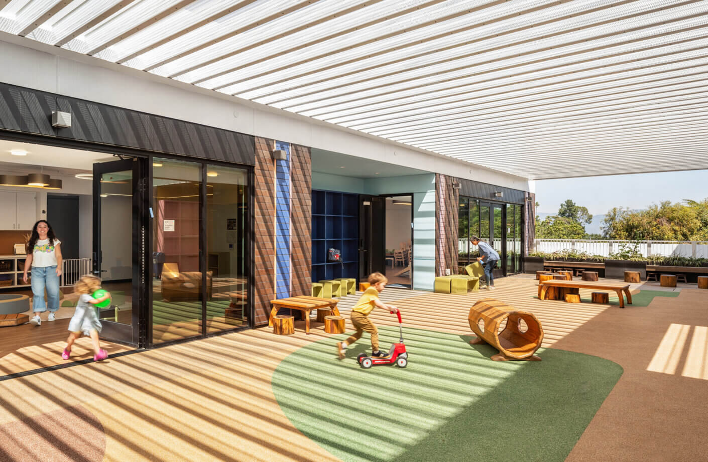 Children play in a vibrant outdoor space with toys and seating, while adults supervise in a well-lit, open area.
