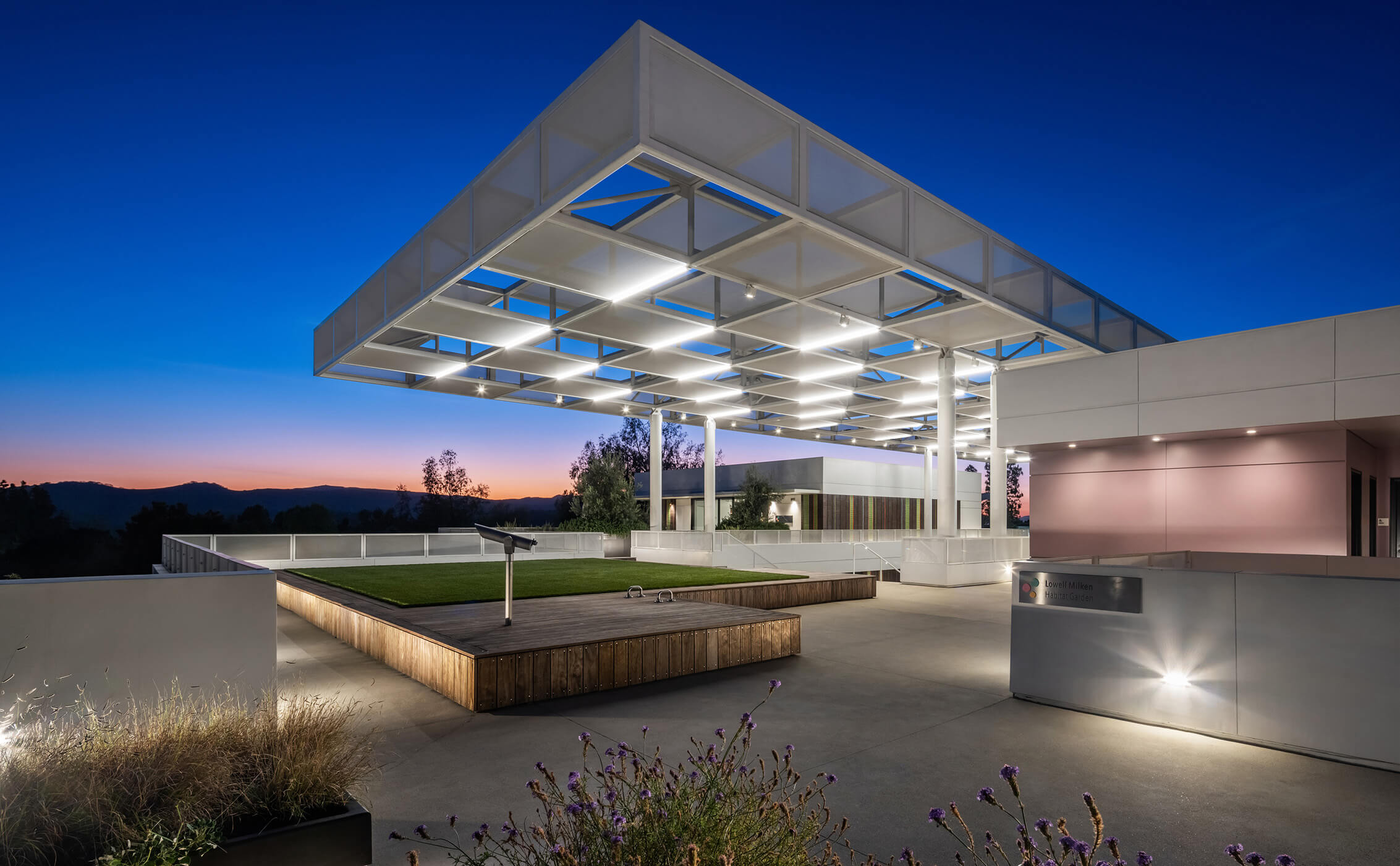 Architectural outdoor space featuring a structured canopy, seating areas, and landscaped greenery at dusk, against a twilight sky.