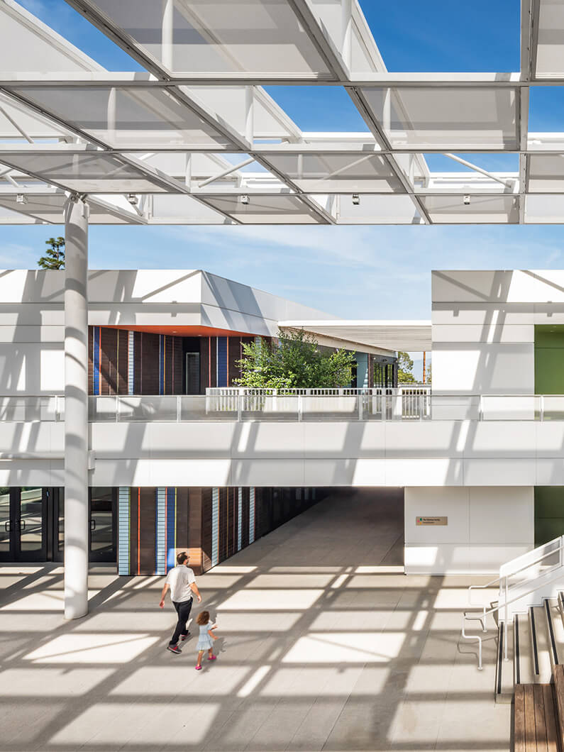 A parent and child walk through an open architectural space with columns, large windows, and geometric shadow patterns on the pavement.