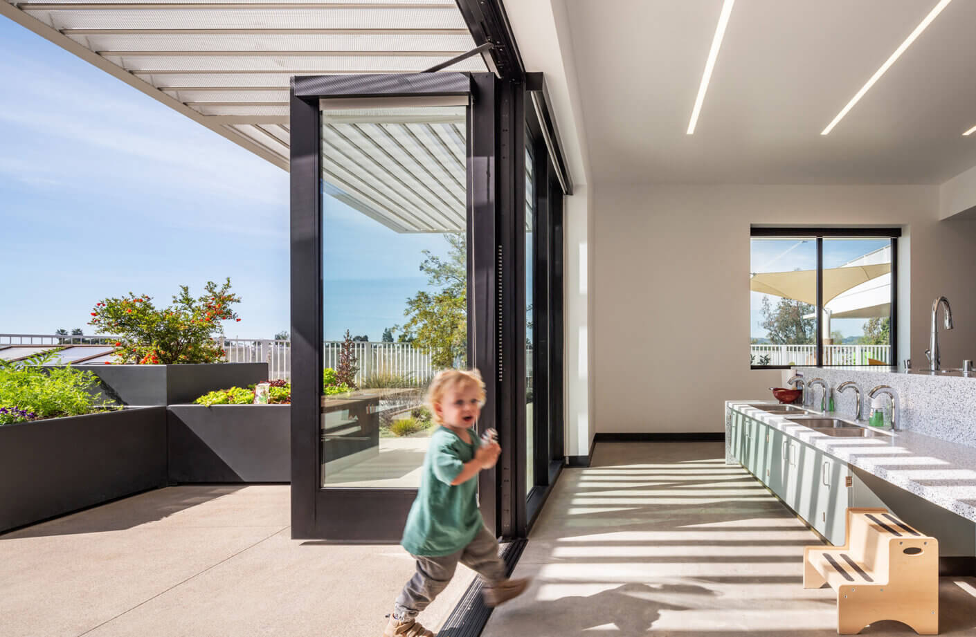 Child running in a spacious kitchen with large glass doors opening to a patio and garden, featuring modern appliances and natural light.