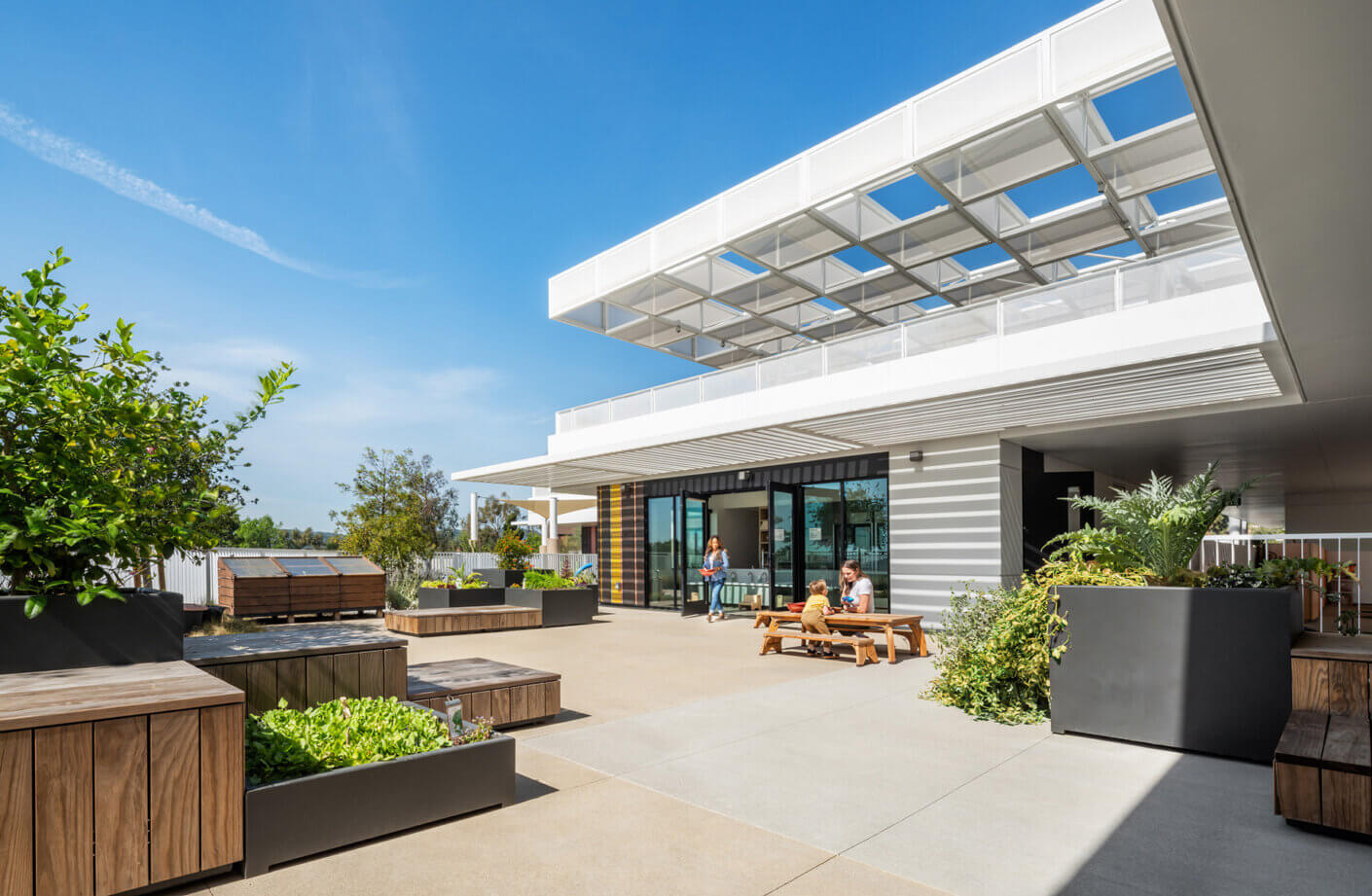 Outdoor space featuring planters and seating, with people enjoying the area in front of a contemporary building under a clear blue sky.