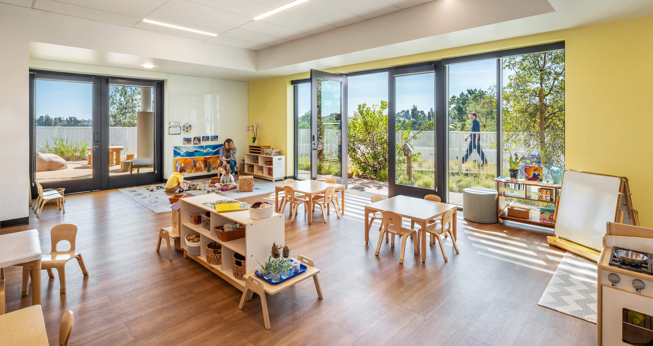 Bright classroom with natural light; children engaging in play. Features wooden furniture, art supplies, and outdoor views through large windows.