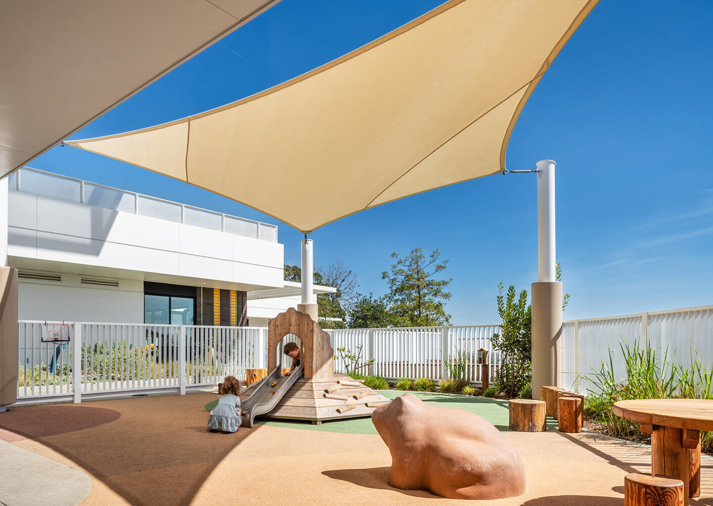 Play area featuring a slide, rock sculpture, and shaded seating. A child interacts with the play structure under a clear blue sky.