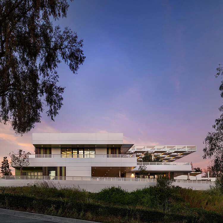 Architectural building with expansive windows and layered design, set against a sunset sky and surrounded by trees and greenery.