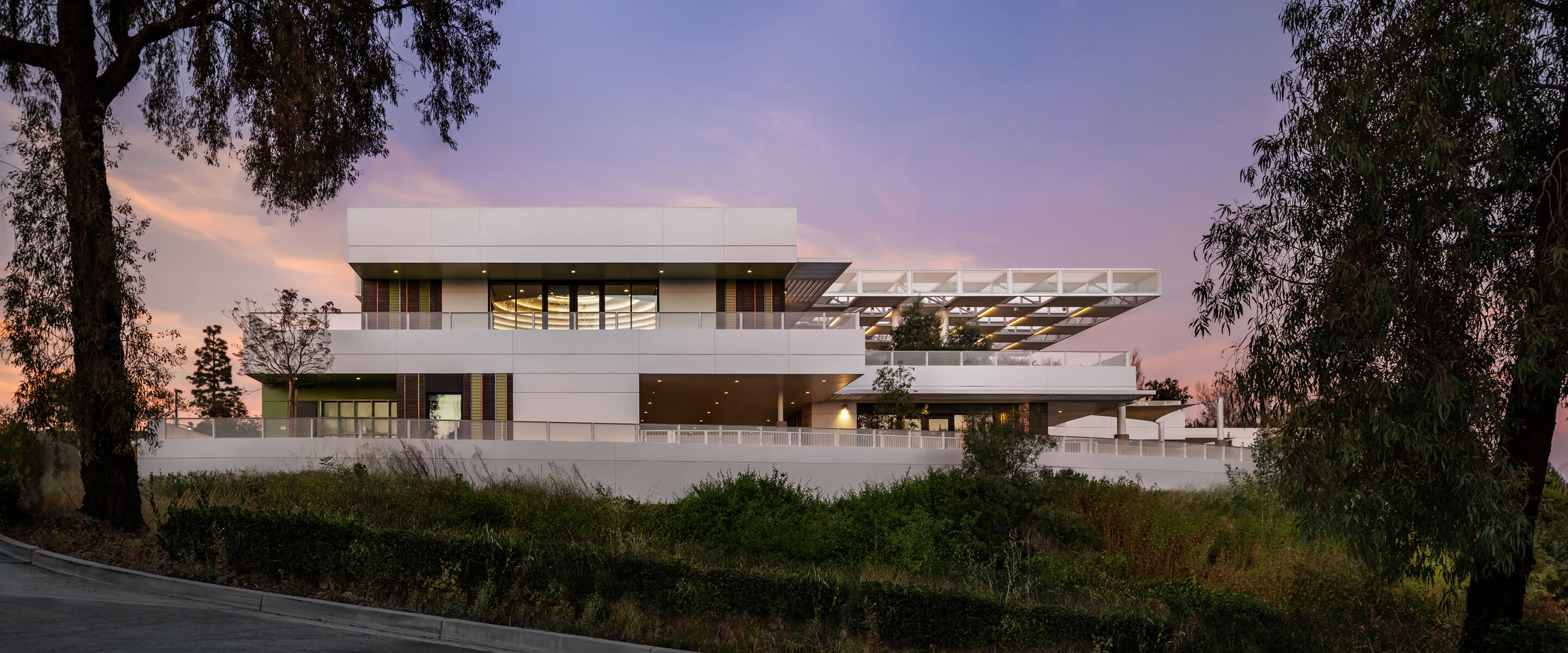 Architectural exterior of a multi-level building surrounded by trees and greenery, captured during sunset with a colorful sky.