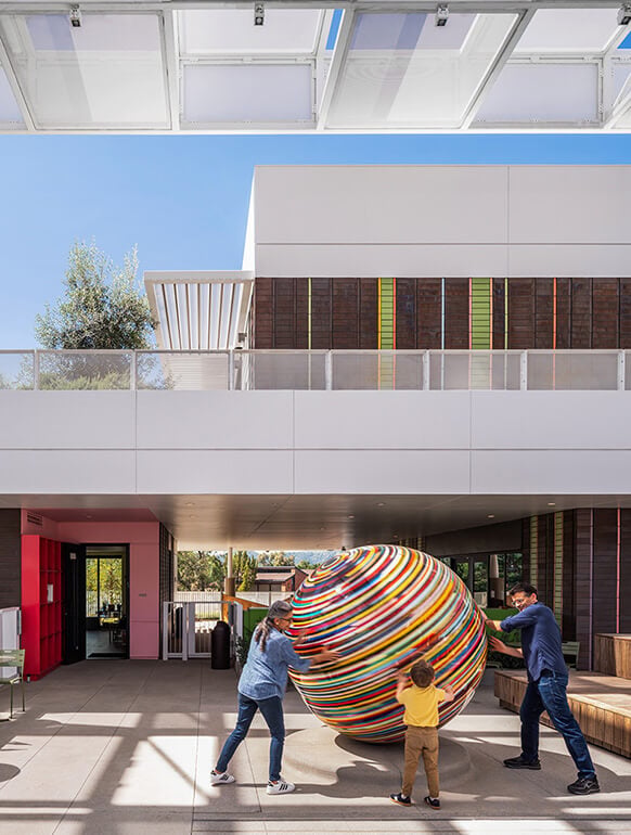 Three people play with a large, colorful sphere in an open architectural space with a sleek design and greenery outside.