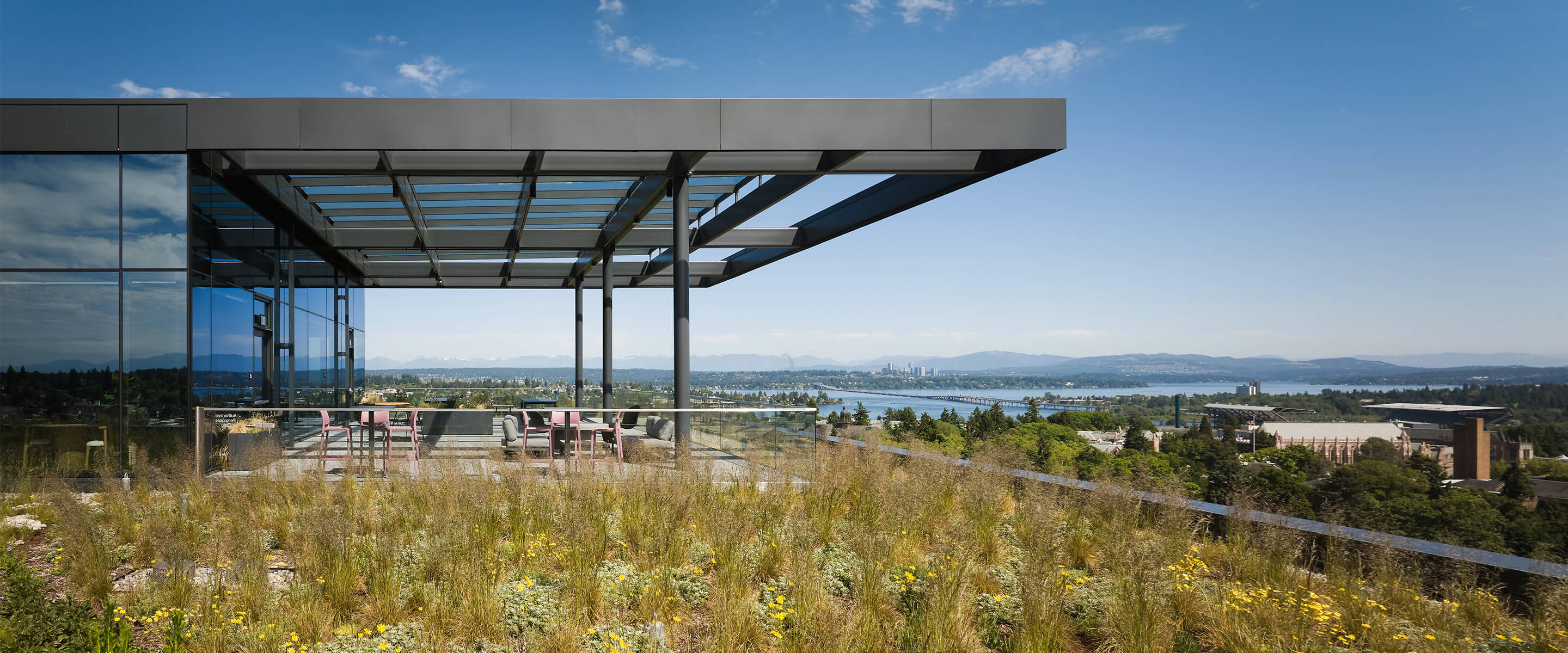 Rooftop patio with glass railings, seating area, and green landscaping overlooking a city and water landscape. Clear blue sky in the background.