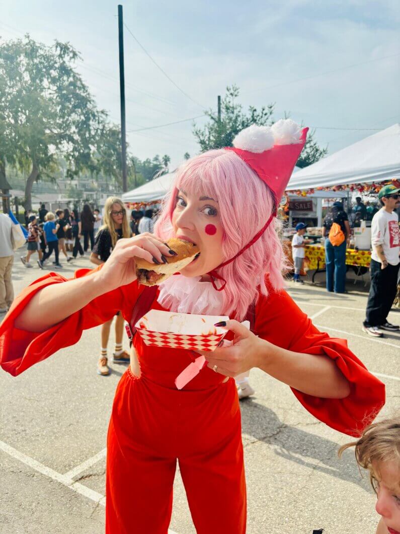 A performer in a red outfit and pink wig enjoys a hot dog at a street festival, surrounded by a lively crowd and food stalls.