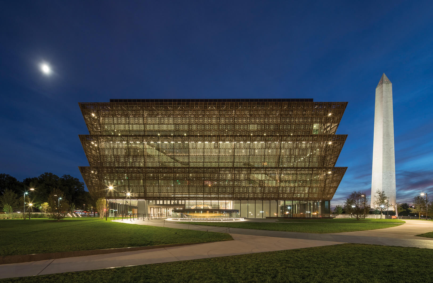 National Museum of African American History and Culture at dusk, showcasing its unique architectural design and lighting, with the Washington Monument nearby.
