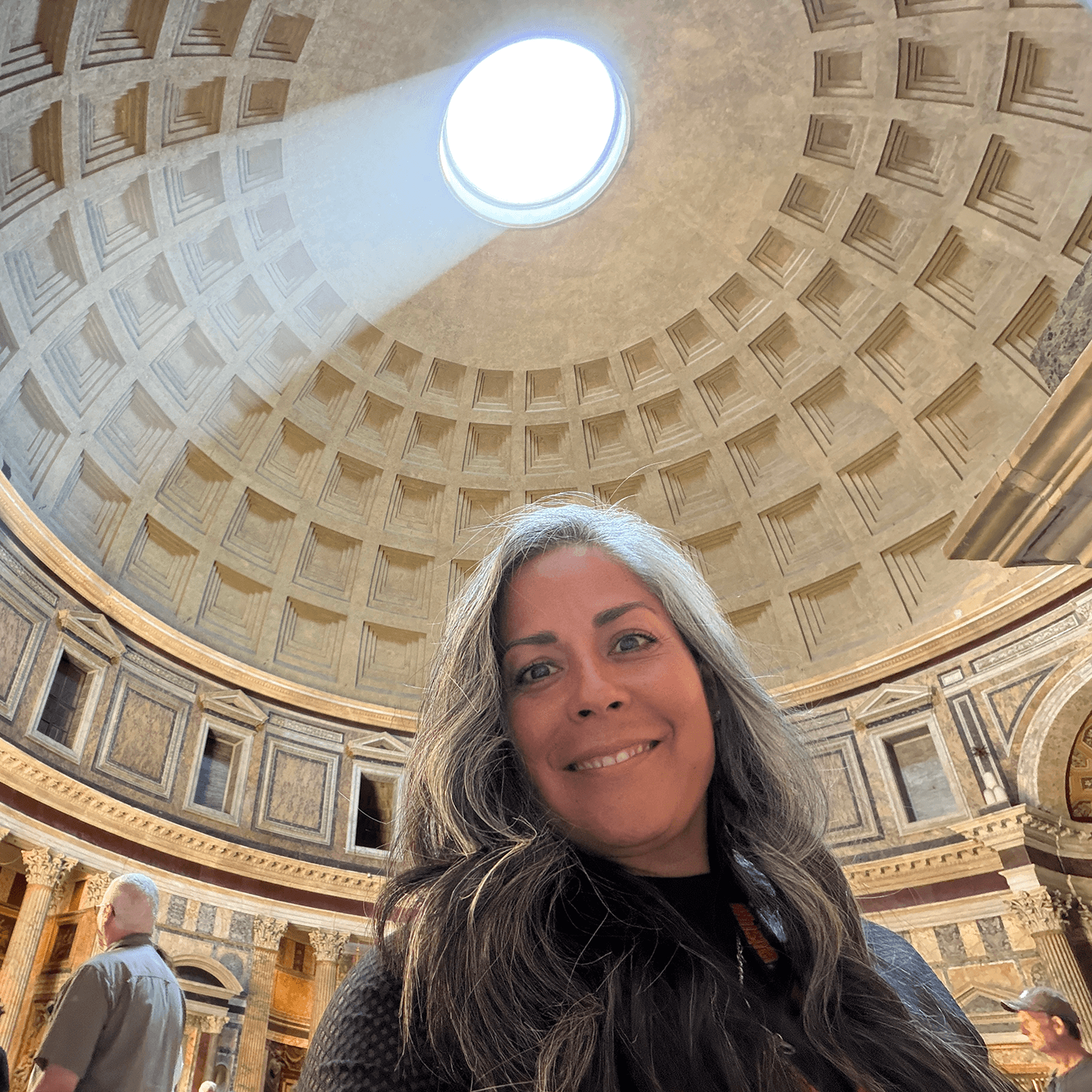 Woman smiling in the Pantheon, featuring a circular skylight and intricate dome design. Visitors explore the architectural details.