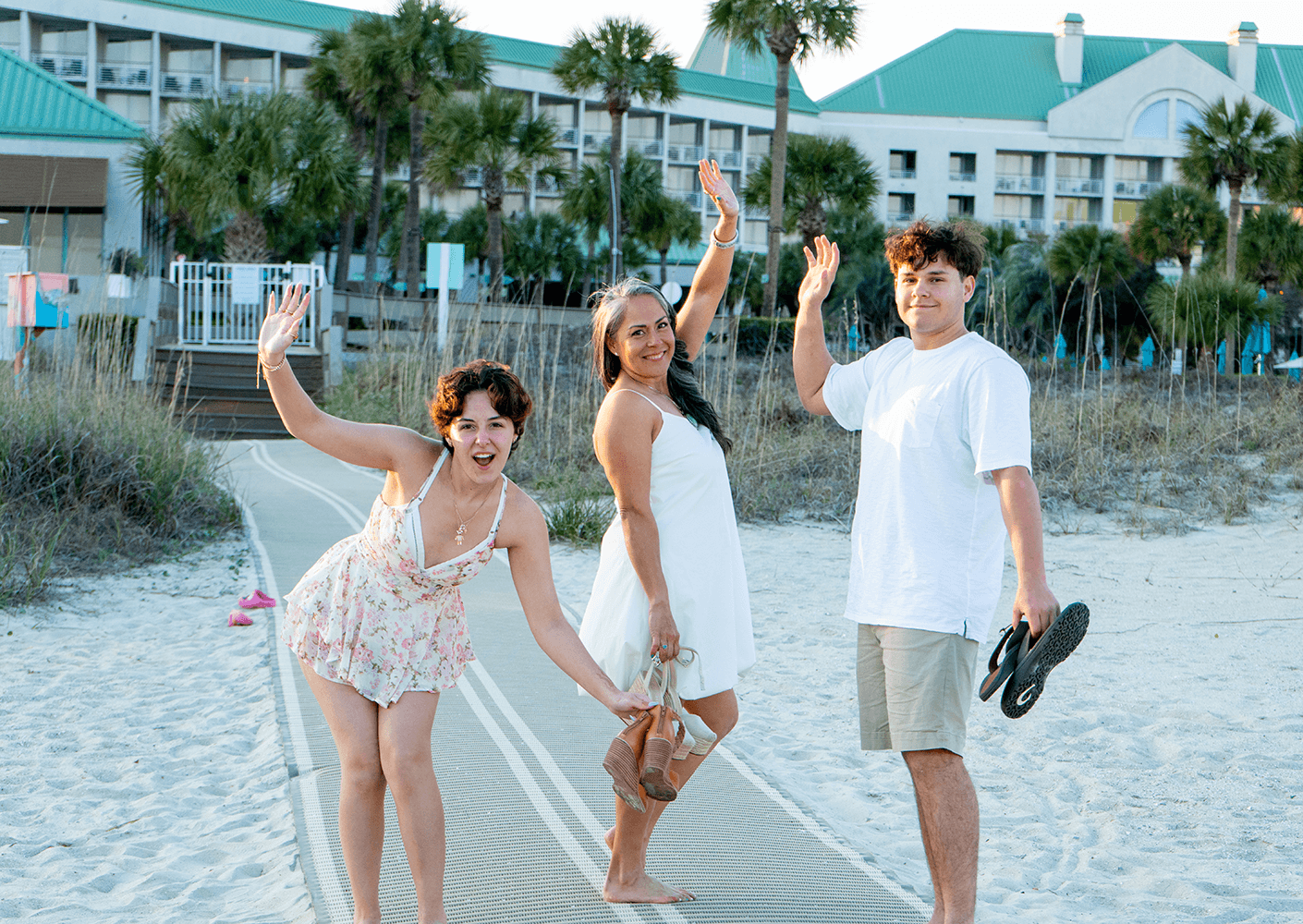 Three people wave happily on a sandy beach path, with palm trees and a hotel in the background during golden hour lighting.