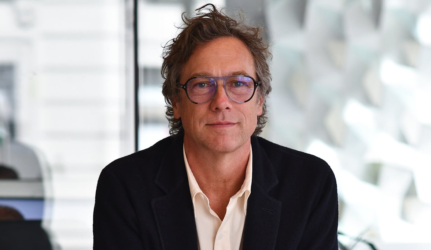 Portrait of a man with curly hair and glasses, seated against a blurred background, with a neutral expression. Professional setting.
