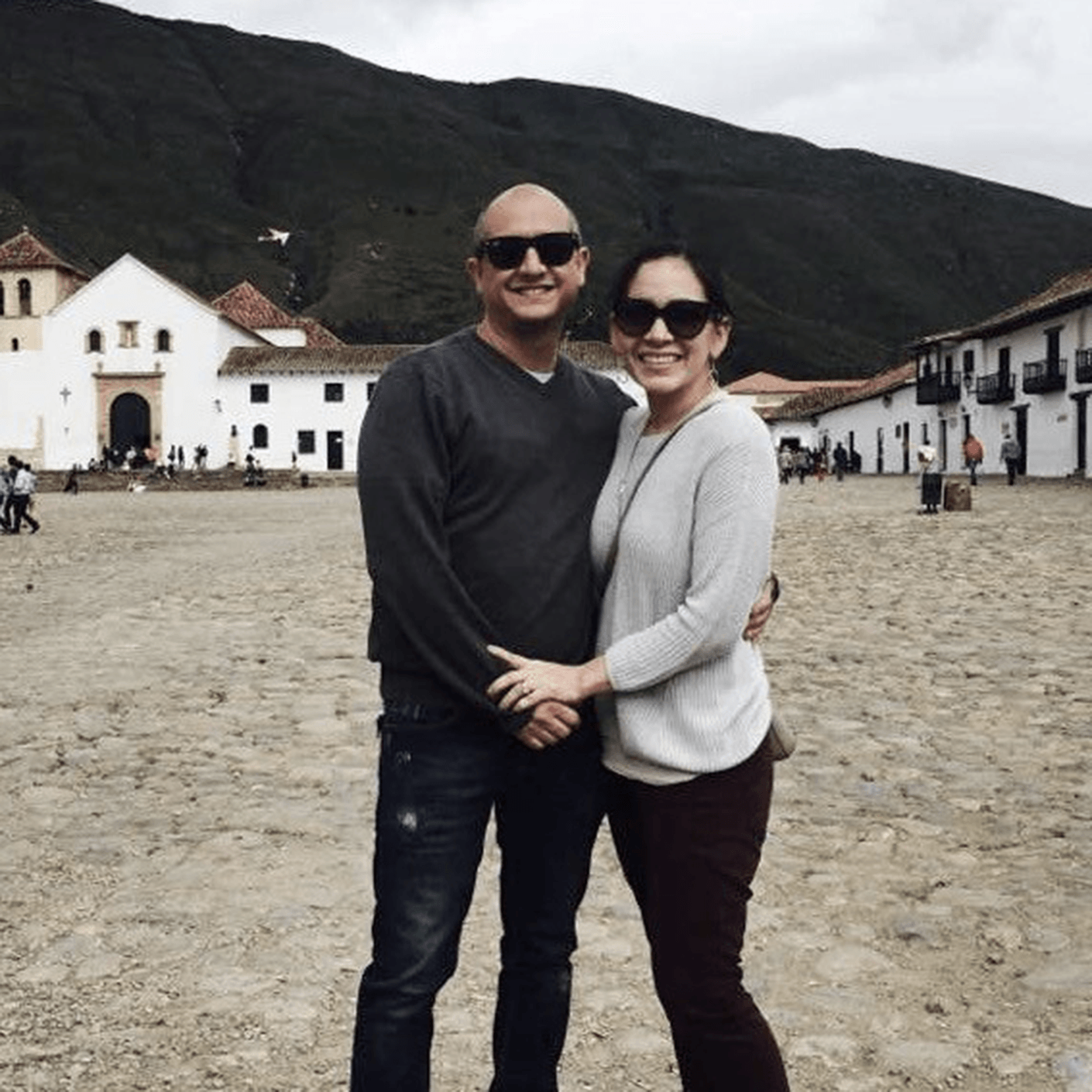 Couple posing together in a cobblestone plaza, with historic buildings and mountains in the background. Overcast sky and casual attire.