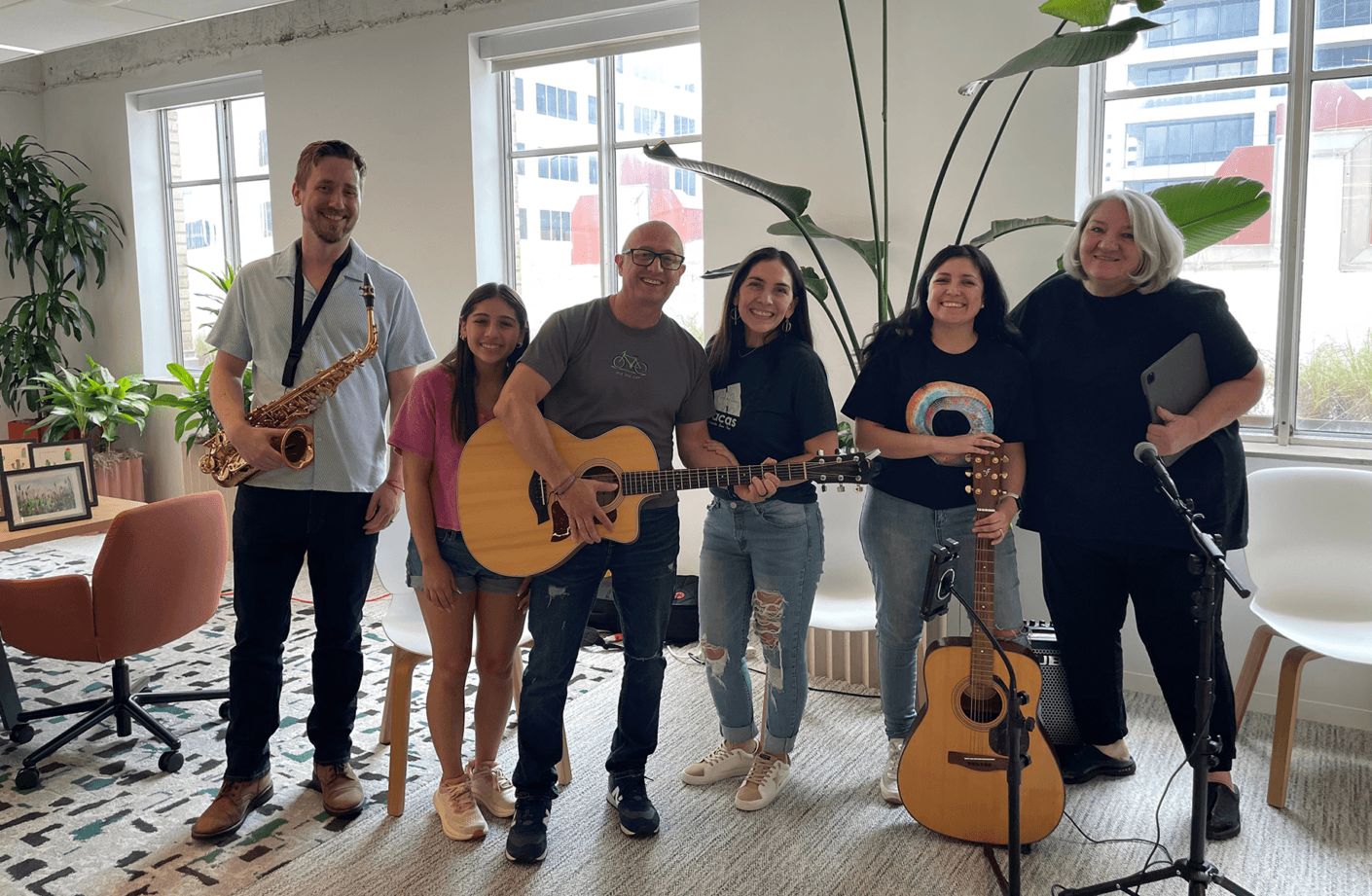 Group of musicians smiling together in a bright indoor space, with instruments including a saxophone and acoustic guitars. Green plants in the background.