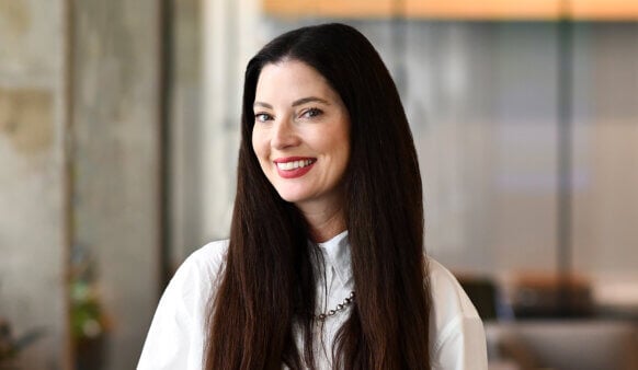 Smiling woman with long dark hair and a white blouse, posed in a well-lit, contemporary office setting.