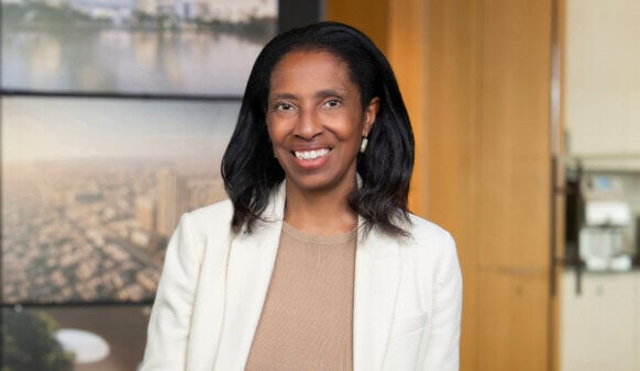 Portrait of a professional woman smiling in a well-lit office setting with a cityscape background. She wears a light blazer and a neutral top.