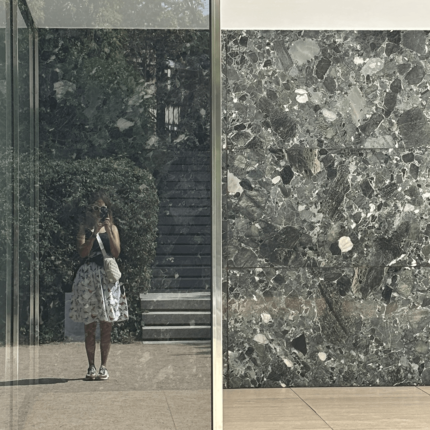 Woman in a patterned dress stands in front of a glass wall reflecting her image. Behind her is a textured stone wall and greenery.