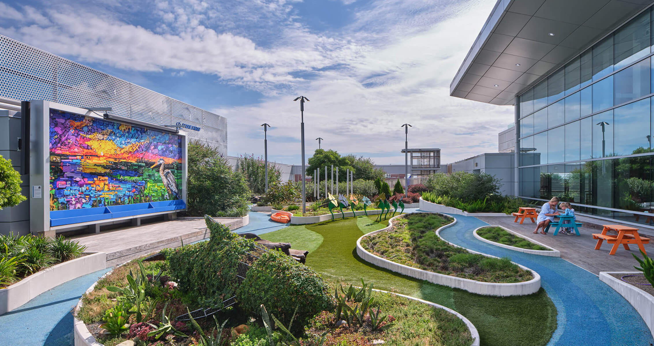 Outdoor space featuring colorful art installation, green landscaping, and seating areas, along with a building with large glass windows.