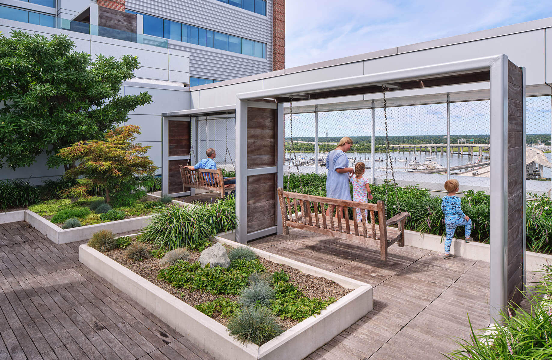 Rooftop garden with swings, featuring a family enjoying the view of a marina and green landscaping in a serene environment.