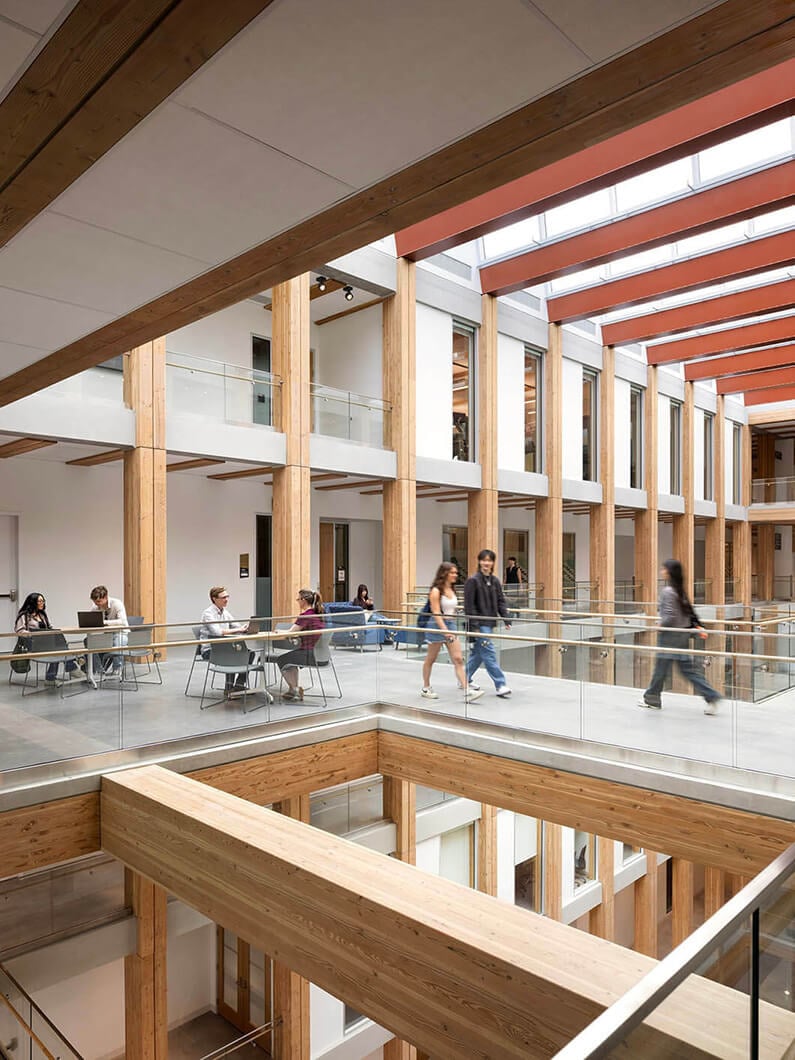 Interior view of a spacious building with wooden beams, glass railings, and people engaging in work and conversation on different levels.