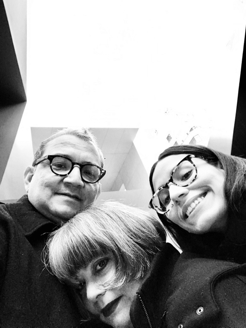 Three people take a close-up selfie, smiling, in a bright, minimalistic indoor space with geometric architectural elements.