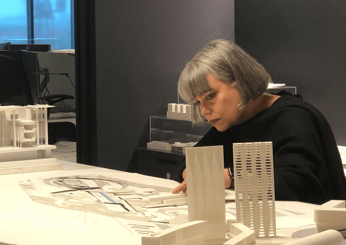 Architect examining a detailed architectural model and plans in an office setting, with white structures present on the table.