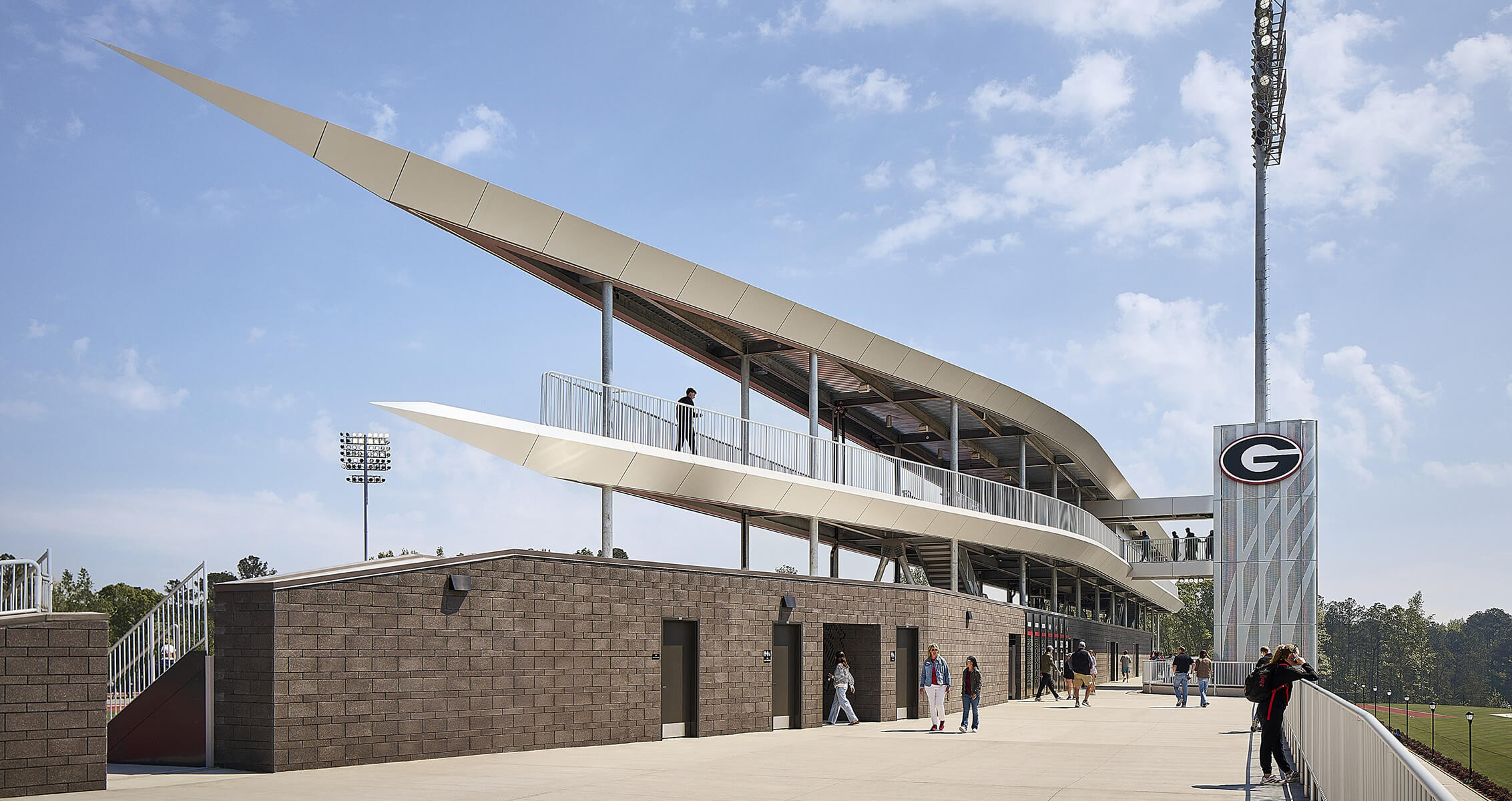 Exterior view of a sports complex with a unique roof design, featuring people accessing the upper level and nearby lighting towers.