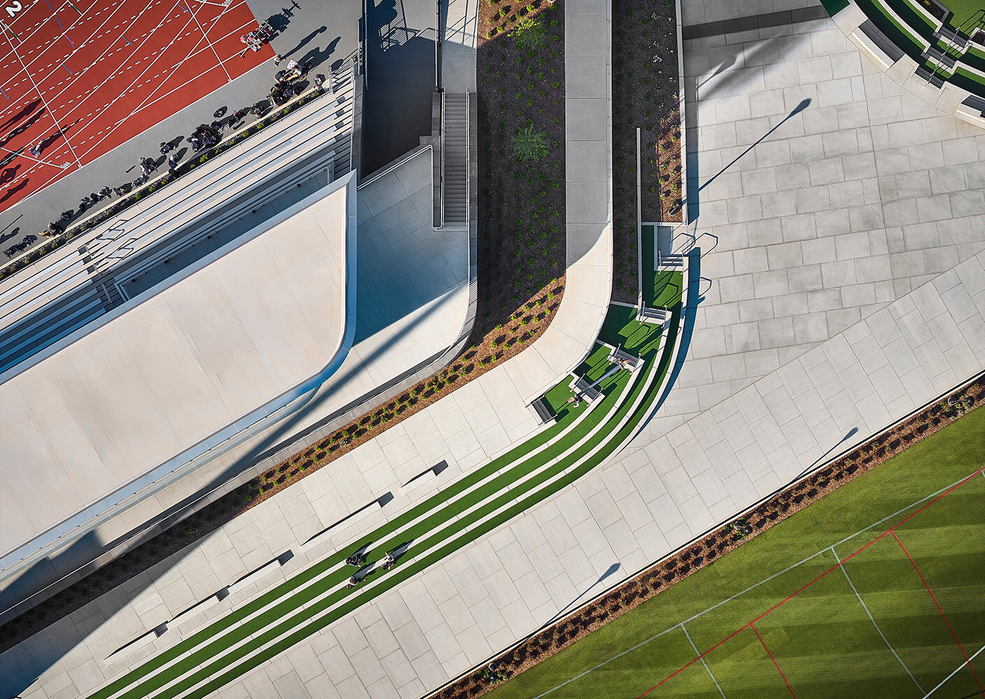 Aerial view of a sports complex featuring a running track, landscaped seating areas, and winding pathways alongside grassy fields.