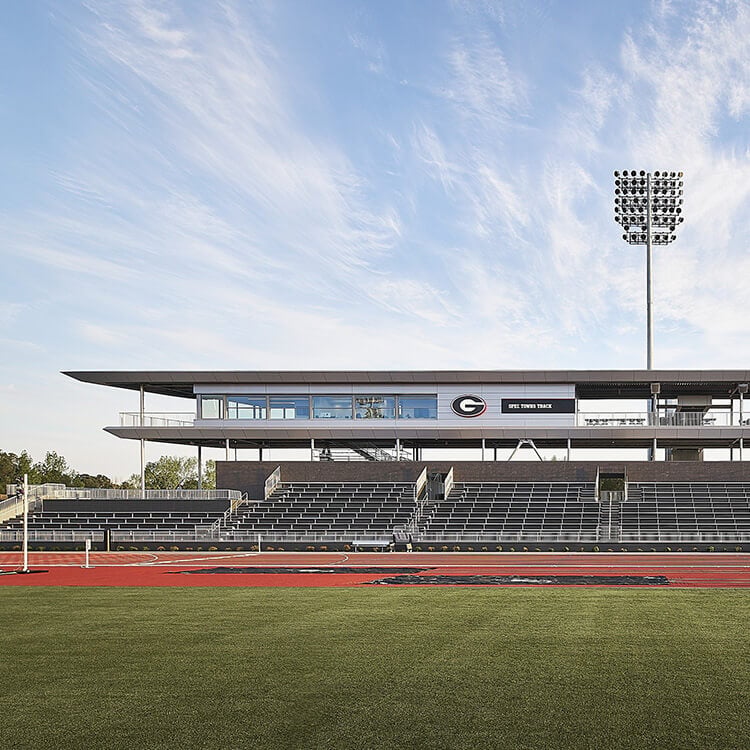 Athletic facility with a grandstand and press box overlooking a track field under a blue sky and stadium lights. Features grass and track surfaces.