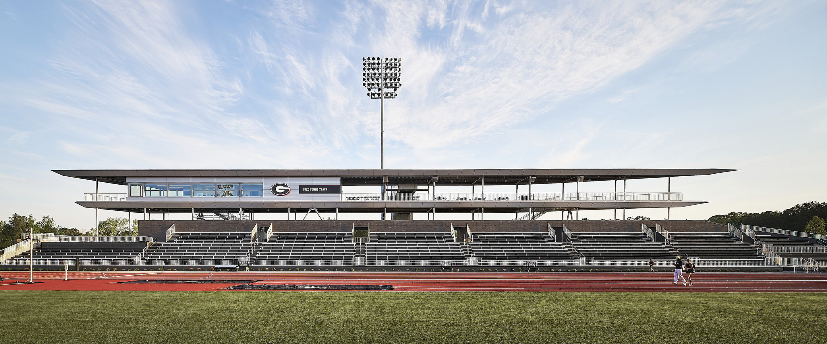 Athletic facility featuring a spacious grandstand and a press box, situated beside a track field under a clear sky.