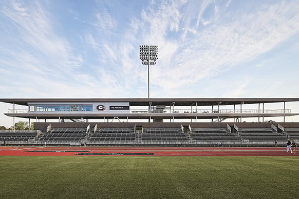 Stadium view showing grandstand seating, press box, and field lights under a clear sky. Track and turf visible in foreground.