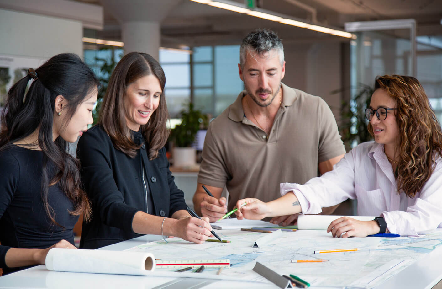 Four professionals collaborate over architectural plans and blueprints on a table, using various tools in a well-lit workspace filled with greenery.