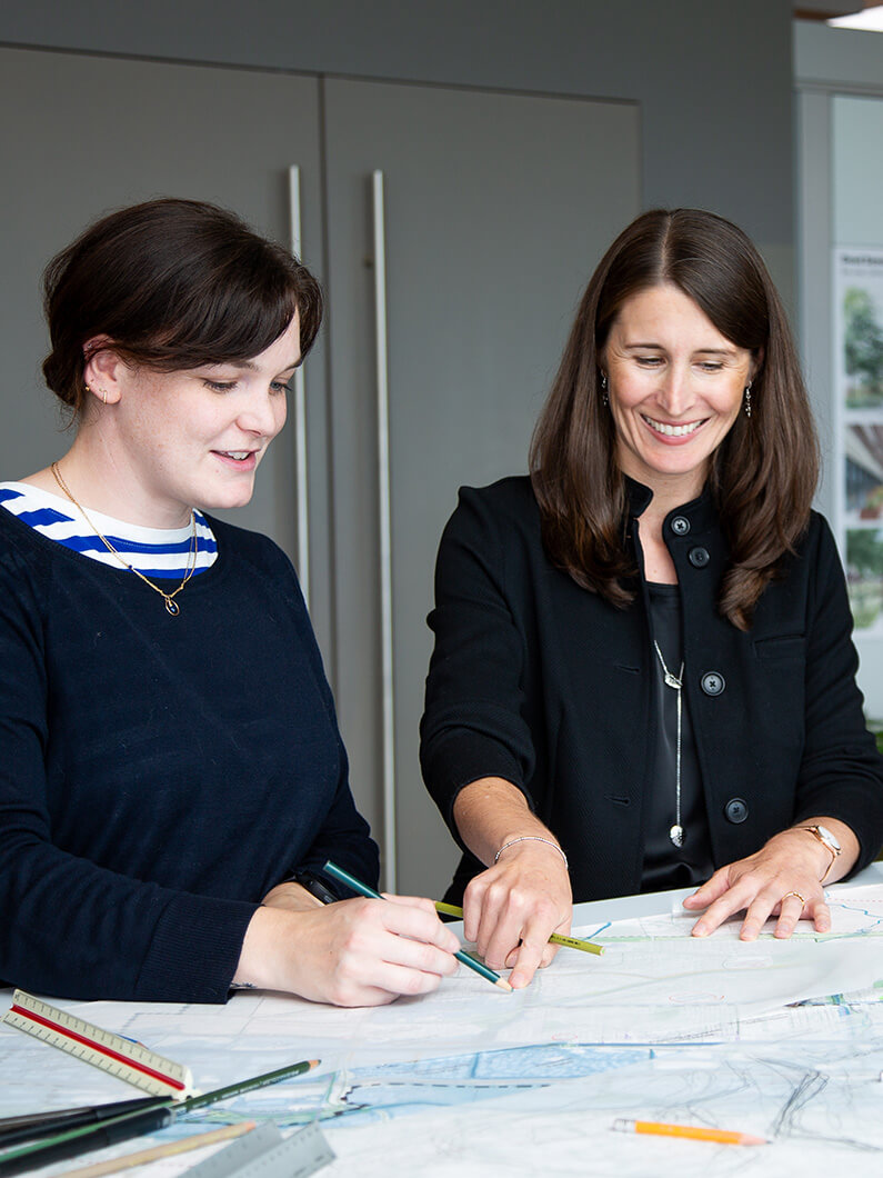Two women collaborate over architectural plans, using pencils and colored pens to sketch and discuss their ideas in a professional setting.