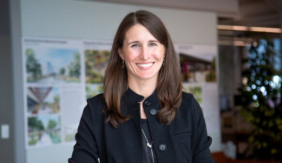 Smiling woman in a black jacket stands in an office, with architectural designs and green plants visible in the background.