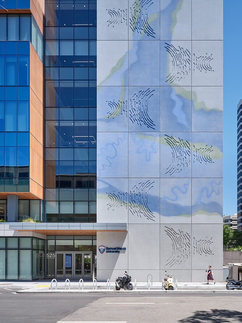 Exterior view of a university building featuring large glass windows and artistic wall designs in blue and green hues. Pedestrians and bikes visible.