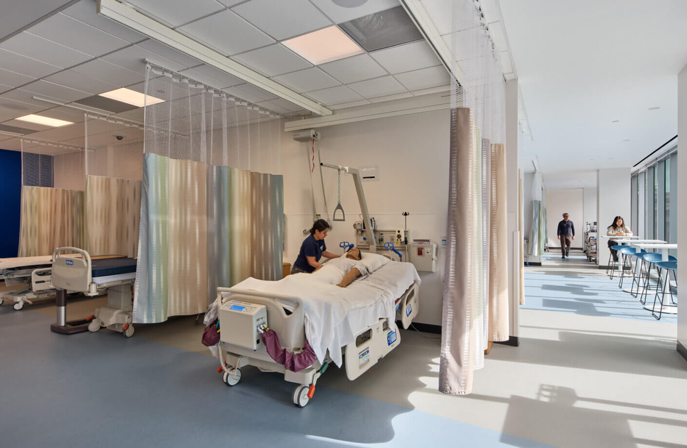 Healthcare facility interior featuring a patient bed, medical staff, and waiting area with privacy curtains and large windows.