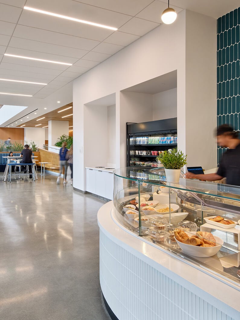 Bright, spacious café with a display case filled with pastries and a beverage fridge against a textured wall. People walk through the area.