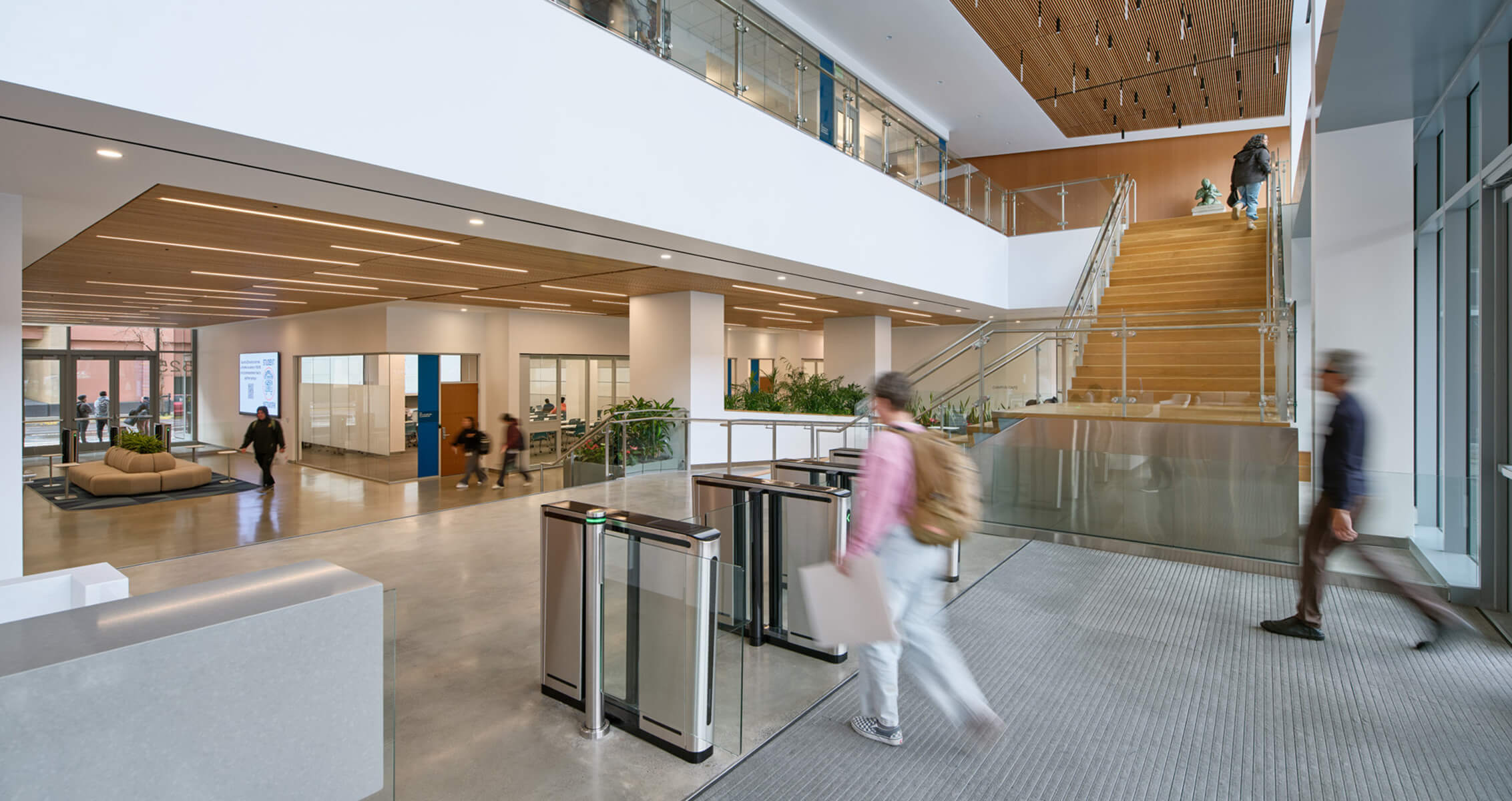 Interior view of a building lobby featuring a seating area, reception desk, glass barriers, and stairs, with people walking through the space.