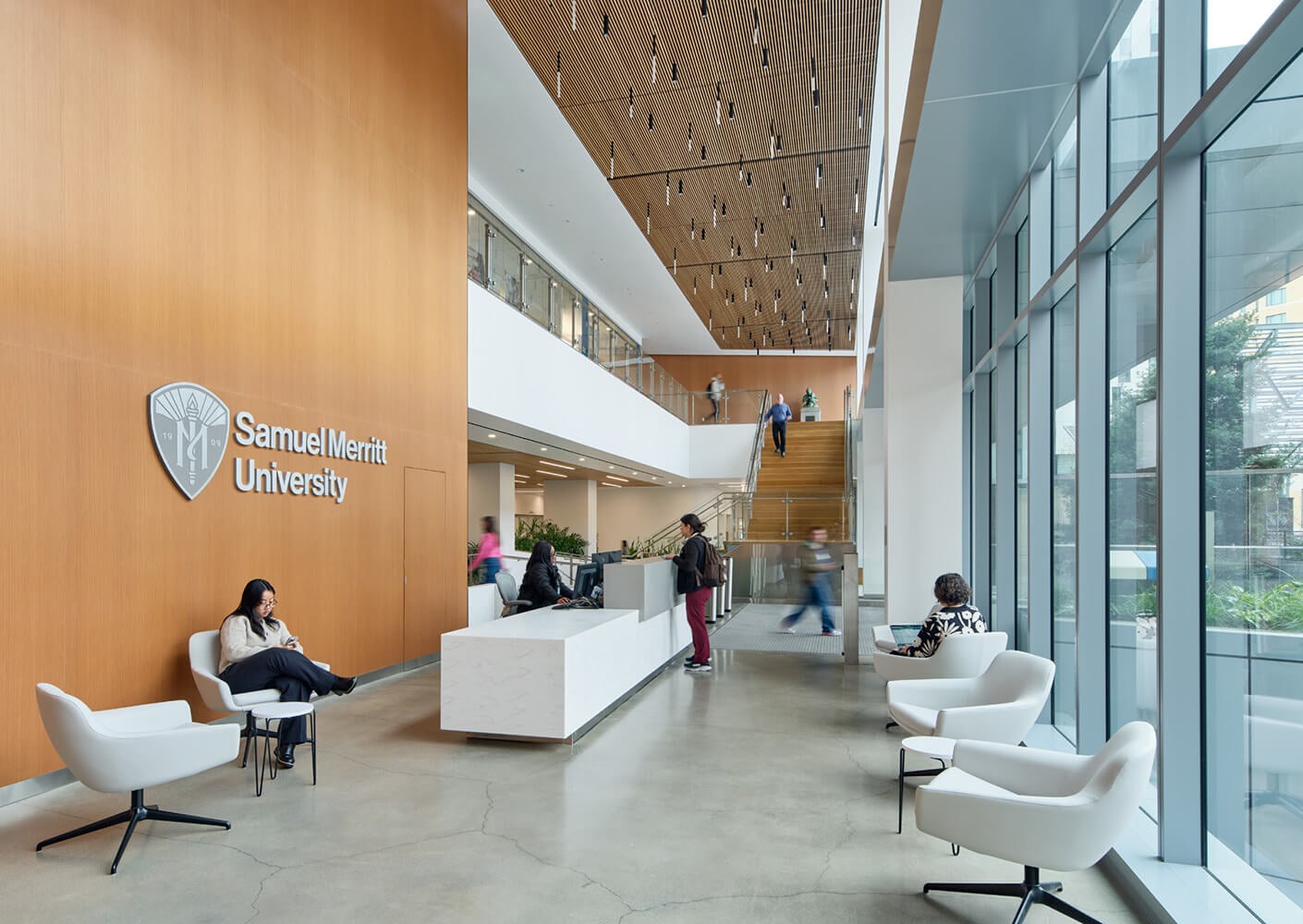 Interior view of Samuel Merritt University, featuring a reception area, seating, and a staircase. Students and visitors interact within the space.