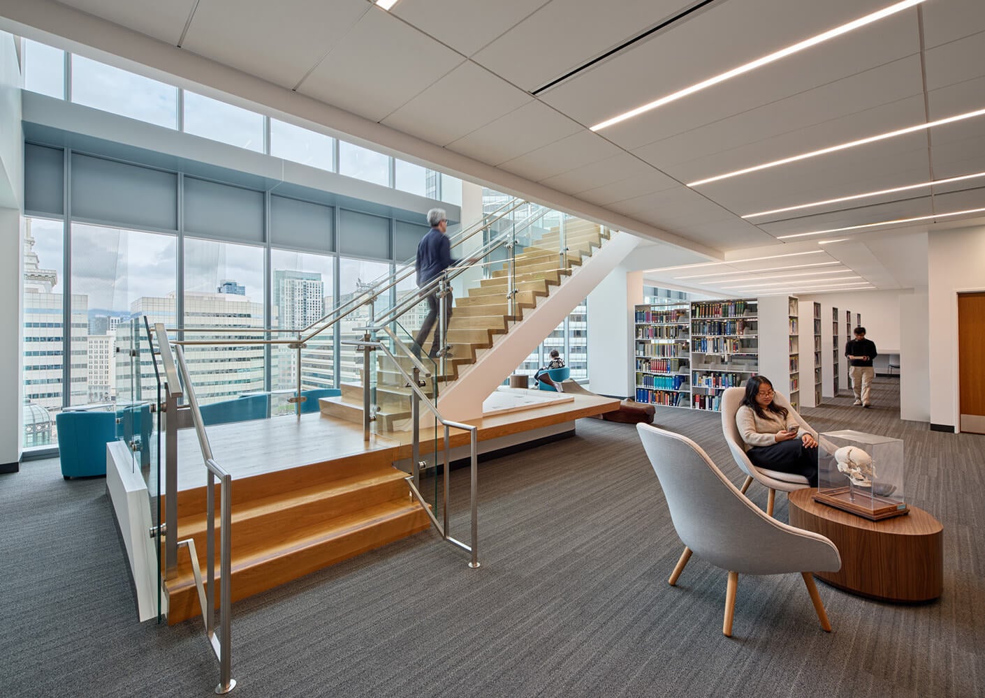 Interior of a library featuring a staircase, seating areas, bookshelves, and large windows with city views. Visitors engage with various spaces.