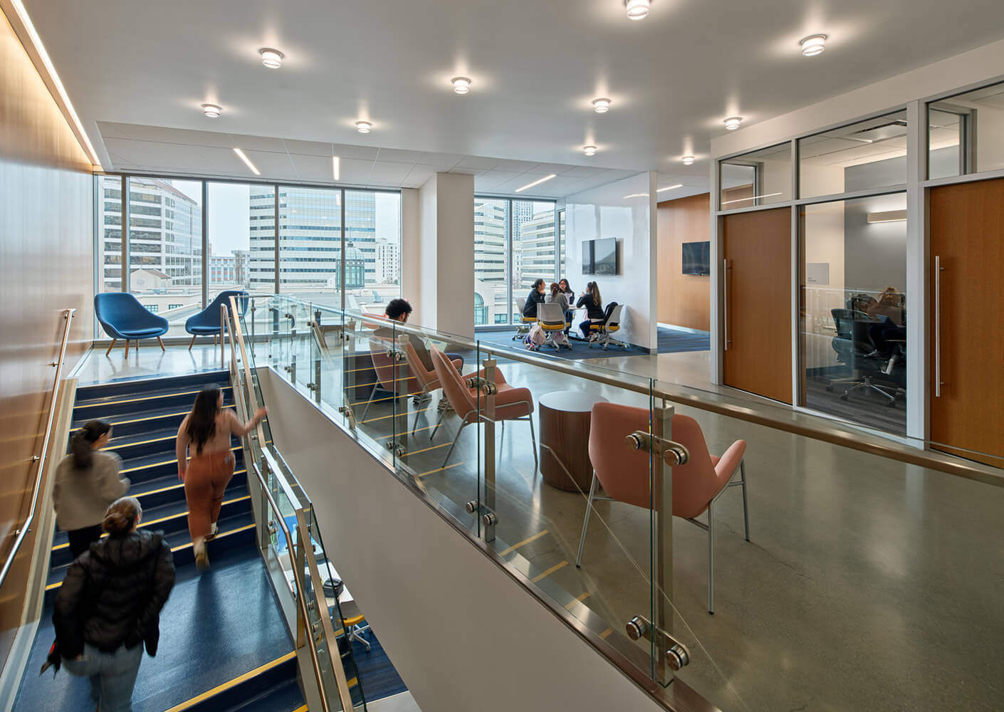 Office interior featuring a staircase, glass railing, and seating areas. People collaborate in a conference room and lounge area with city views.
