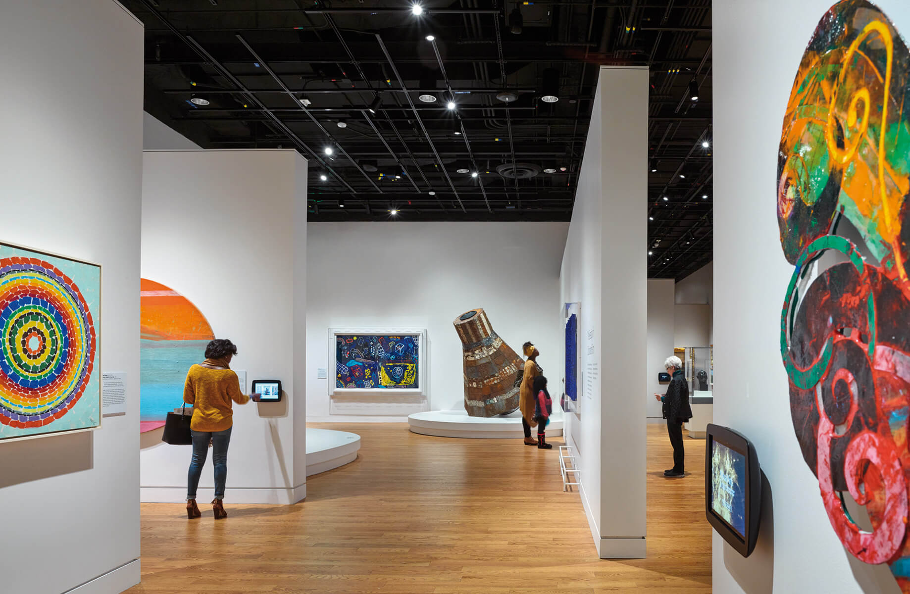 Art gallery interior featuring colorful paintings, sculptures, and visitors engaging with the exhibits. Wooden flooring and bright lighting enhance the space.