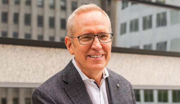 Portrait of a smiling man wearing glasses and a blazer, standing outdoors near a building with reflective glass.